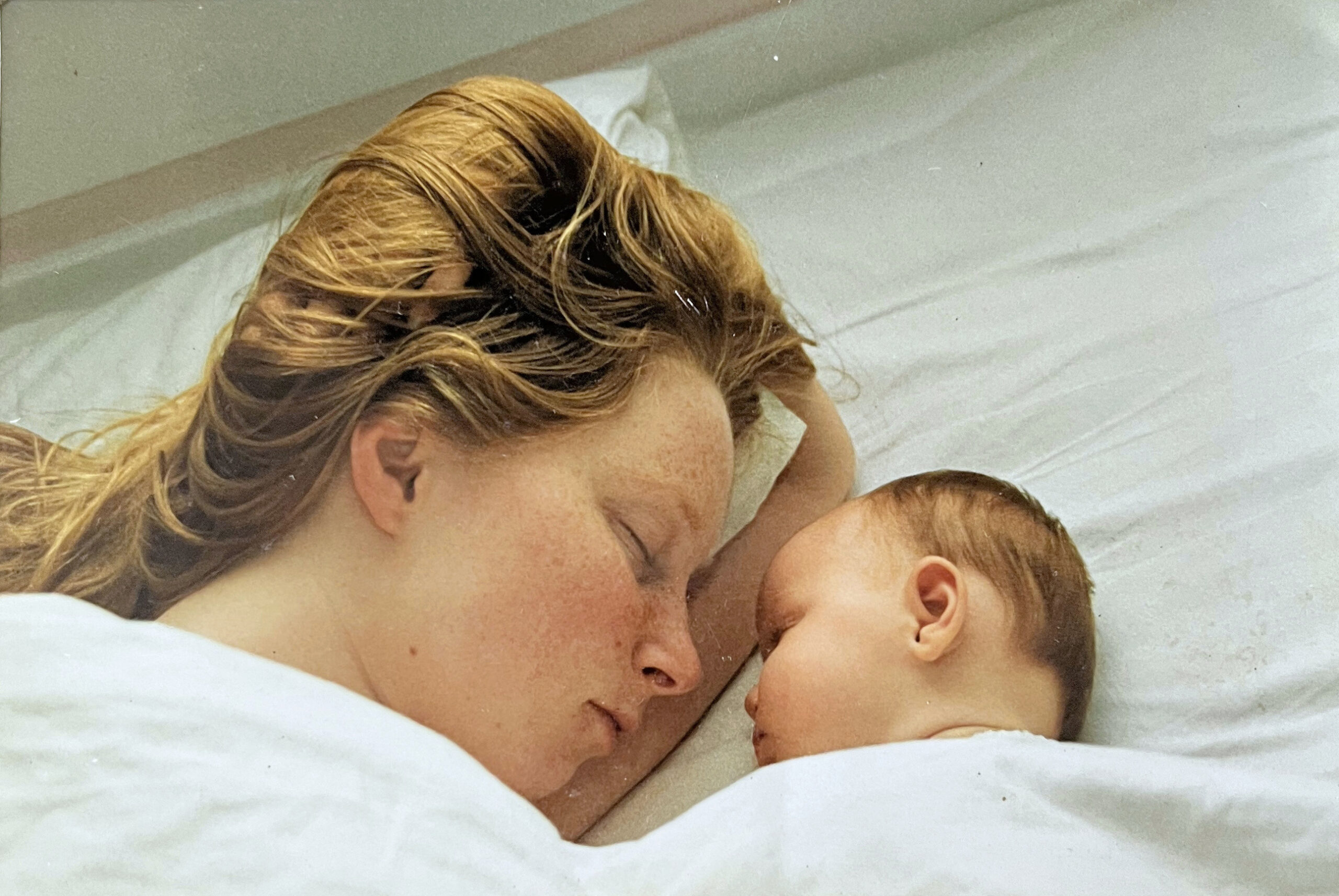 A mother and her baby fast asleep in the family bed, facing each other, their heads together.